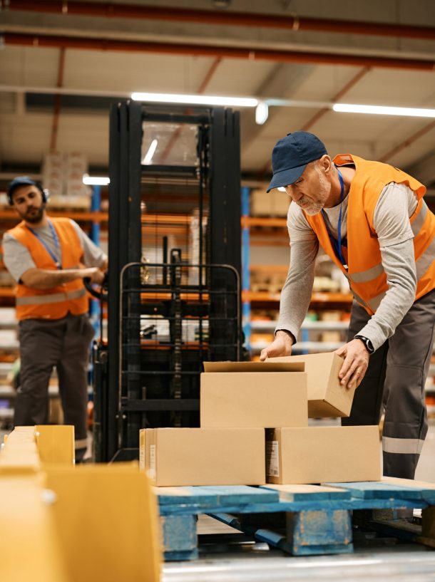 Mature worker stacking cardboard boxes while working with coworker at distribution warehouse.